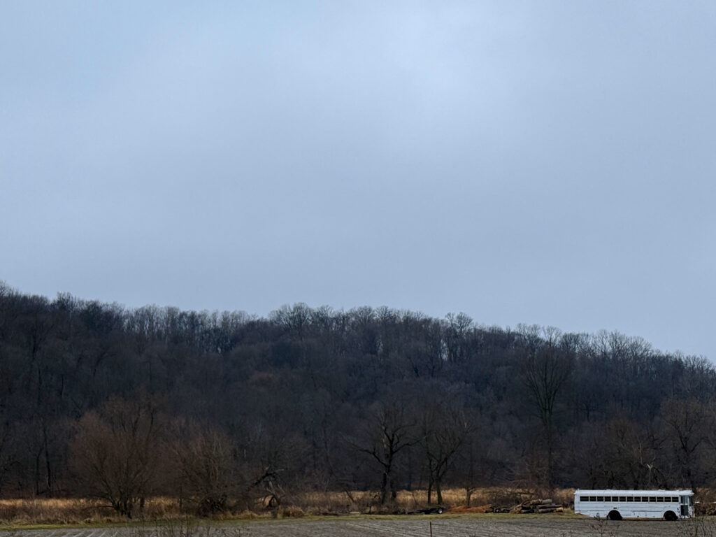 A black and white photograph of an open field backdropped by a forested hill in later winter. A bus sits in the right-hand foreground. This image links to the page "Cream Soda and the Loch Ness Monster"